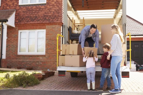 A professional house removal scene in a suburban residential area shows a Croydon Man with Van team member, clad in casual attire, loading large cardboard boxes into the back of a moving van parked on a paved driveway. The van's interior reveals partially packed furniture and belongings, with wooden furniture pieces visible inside the open cargo area. A family, including a woman in a striped top and jeans, along with two children—a girl and a boy—are standing on the driveway, overseeing the process. The children, a girl and boy, are dressed casually and are reaching up towards the man, possibly handing or receiving items. The background features a red brick house with white-framed windows, a well-maintained garden bed with some greenery, and neighboring houses with similar brick exteriors and tiled roofs. The scene is illuminated by natural daylight, emphasizing the neat, organized preparation for a home relocation in Croydon or nearby areas in South London, highlighting the professional and careful approach of Croydon Man with Van in house removals and safe transportation.