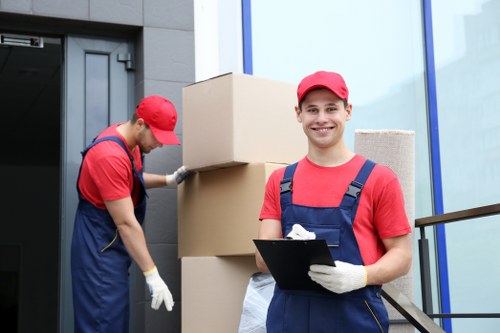 Two professional removals workers dressed in red caps and blue uniforms are engaged in a house removal process outside a modern residential building. The worker on the left is lifting a large cardboard moving box, standing next to stacked boxes arranged vertically against the exterior wall, which appears to be made of grey siding or cladding. The other worker, positioned to the right, is smiling at the camera and holding a clipboard, indicating supervision or organization of the move. The background features a contemporary house with glass windows and a dark-framed entrance door, suggesting a residential property in Croydon or nearby in the postcode area. The environment is well-lit with natural daylight, highlighting the clean, orderly scene of packaging and transport preparations typical of professional house removals services. The image captures the essence of a systematic move, with emphasis on the sturdy, cardboard boxes and the meticulous staff organization, reflecting Croydon Man with Van’s house relocation expertise.