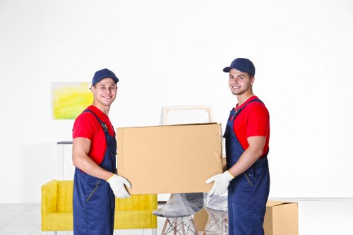 Two professional house removals workers dressed in red t-shirts, blue overalls, and caps stand indoors in a bright, minimalist living room with white walls and flooring. They are smiling and holding a large cardboard box together, which is positioned in front of a small, round glass table with a grey cloth cover. In the background, there is a yellow upholstered sofa and a white side table with a piece of abstract yellow and white wall art above it. The room appears to be part of a residential property, likely in Croydon or nearby in South London, during daytime with natural light illuminating the space. The workers are wearing white gloves, and the scene depicts a typical moment of loading or unloading during a home move, emphasizing careful handling of household items such as boxes and furniture, consistent with house removals services by Croydon Man with Van.