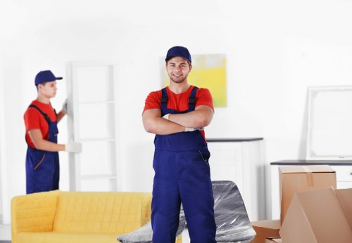 A professional mover from Croydon Man with Van stands confidently in a well-lit room during a house removal, wearing a navy blue cap and matching overalls over a red T-shirt. The individual has arms crossed and a friendly expression. In the background, another mover, also in red and blue work attire with a navy cap, is handling a white shelving unit, possibly preparing to carry or position it. The room features a yellow fabric sofa situated in the foreground, with stacked moving boxes in cardboard and plastic wrapping nearby. The interior has a white paneled wall, a dark countertop, and a large window that allows natural light to illuminate the space. The scene captures the organized movement of household items typical of a house removal in Croydon, with furniture, boxes, and packing materials arranged systematically to facilitate a smooth relocation process, illustrating the professional service provided by Croydon Man with Van.