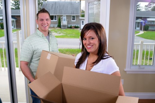 Inside a bright, well-lit residential house, a man and a woman are engaged in a house removal process, with the woman in the foreground holding an open cardboard box. The woman has shoulder-length brown hair and is wearing a white top, smiling as she looks towards the camera. Behind her, the man, dressed in a light green, short-sleeved, plaid shirt, also smiles and is standing beside her, holding a similar box. The background shows a view of a street through large windows, revealing a suburban neighbourhood with green lawns, detached houses, and a quiet residential street. Inside the room, the walls are painted in a light, neutral colour, and the space appears organized, ready for moving activities. The boxes are plain brown, made of cardboard, and vary in size, indicating they are used for packing personal belongings during a house move. This scene emphasizes preparations for a residential move, typical of services provided by Croydon Man with Van, a local house removals company serving the Croydon area near the postcode CR0.