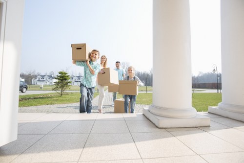 Four individuals are standing outdoors on the steps of a building with large white columns, engaged in a house removal activity. The woman in the foreground, wearing a green and white checkered shirt, is smiling and holding a cardboard box above her head, demonstrating a cheerful attitude. Behind her, three other people, two women and one man, are also carrying cardboard boxes, preparing for loading or unloading. The boxes are plain brown cardboard, some with visible tape, indicating they are packed for a move. The scene takes place in a suburban area with a grassy lawn, small trees, and residential houses visible in the background under a clear sky, suggesting a daylight setting. The building has a stone or concrete tiled floor, and the scene captures the moment of different people actively involved in moving items, possibly part of a local house removal service in Croydon or nearby. The overall environment reflects a typical community surroundings with a professional yet casual atmosphere, aligned with house removals operations carried out by Croydon Man with Van.