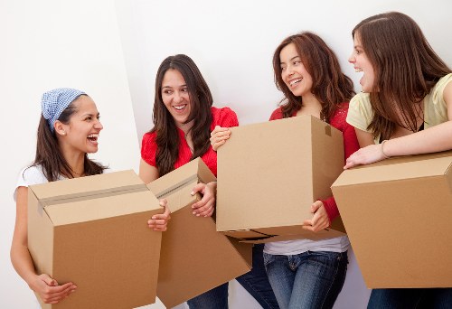 Four women with light skin, smiling and engaging in conversation, are holding large cardboard boxes filled with household items, including a small wooden table and a fabric-covered chair in an interior space with white walls. The women are casually dressed, with one wearing a blue headscarf, another in a red top, and the others in colorful tops. The scene appears to be in a residential setting, possibly a hallway or room within a house in Croydon, during a house move or relocation process, with the focus on packing or unpacking belongings in preparation for a removal service by Croydon Man with Van. Bright, natural lighting illuminates the space, highlighting the textures of the cardboard boxes and the variety of household objects being carried or handed over. The overall atmosphere suggests efficiency and teamwork in a domestic moving context, subtly supporting local house removals, recycling, and sustainability themes relevant to Croydon postcodes.