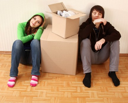 A young woman and a young man with light skin are seated on a wooden floor in a residential setting, suggesting an indoor environment typical of a house in Croydon. The woman, positioned on the left, is wearing a bright green hoodie and blue jeans, with pink and white striped socks; she rests her head on her hand, appearing contemplative or tired. In front of her is a large beige cardboard box filled with packing paper, placed on top of a similarly large cardboard box. The young man, seated to the right, is dressed in a black hoodie and grey trousers, with black socks; he has a thoughtful expression, with one arm resting on his knee and his hand near his face. Behind the boxes, the background features a plain, lightly textured pale wall, indicative of a typical home interior in the Croydon area. The scene captures a moment of packing or unpacking, characteristic of house removals or relocation activities, with natural lighting filling the room, emphasizing the textures and colors of the household items and clothing.