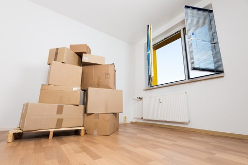 A room with white walls and a light wooden floor shows a stack of cardboard moving boxes of various sizes, some sealed with tape, placed on a wooden pallet in the left foreground. The boxes appear new and ready for a house move, containing personal belongings or household items. On the right side, a large window with open blue Venetian blinds allows natural light into the space, revealing a residential street view outside. Below the window, a white radiator is mounted on the wall. The room appears empty apart from the boxes, suggesting a moving or packing process. The environment looks clean and tidy, with minimal furnishings, indicating preparation for house removals in Croydon or nearby areas. The setting captures an interior scene typical of an in-progress move, with simple, modern decor and a focus on the packing activity associated with house relocation services by Croydon Man with Van.