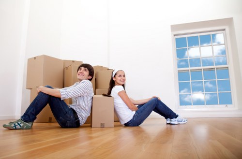 A young man and a young woman with light skin are sitting on a polished wooden floor inside a residential property, leaning against a stack of several cardboard moving boxes of various sizes, which are arranged in a semi-structured manner in front of and beside them. The man, wearing a striped shirt and jeans, is positioned on the left side and smiling, while the woman, dressed in a white top and jeans, is on the right side, also smiling. Behind them, a large double-glazed window with a white frame allows natural daylight into the room, revealing a clear blue sky with some clouds outside. The room has white walls and a minimalistic appearance, typical of a home preparing for a house move in Croydon, with no other furniture or items visible. The setting indicates a house removal or relocation process, consistent with services offered by Croydon Man with Van, demonstrating a cheerful moment of packing or unpacking within a residential property in the Croydon area.