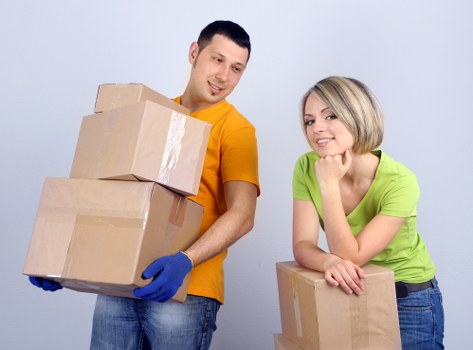 The image shows a man and a woman with light skin, both smiling, in a well-lit indoor setting with a plain light grey background. The man, wearing a bright orange t-shirt and blue latex gloves, is holding three cardboard boxes stacked one on top of the other, with the boxes appearing to be new and of medium size, featuring brown kraft paper and clear packing tape along the edges. The woman, dressed in a lively green t-shirt, is leaning on a small cardboard box resting on a flat surface, with her arms crossed over it and her chin resting on her hands, displaying a friendly expression. Behind them, a partially visible white wall and a faint floor suggest a residential interior, consistent with house remodelling or removals context. The scene reflects a professional house removals service, with the items arranged clearly to show typical moving supplies, such as the cardboard boxes used for packing personal belongings in a house clearance or relocation within Croydon or nearby areas in the UK, as suggested by the image's professional styling and content.