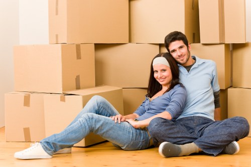 A young couple sitting on a wooden floor in a residential environment surrounded by numerous large cardboard boxes, which are sealed with packing tape and vary in size. The woman, wearing a light blue top and jeans, is leaning back against the man, who is dressed in a light blue shirt and dark jeans, with both smiling at the camera. Behind them, there is an indoor setting with a plain wall, suggesting a house or flat interior. The scene depicts a typical house removals or moving process in Croydon, with the couple appearing relaxed and prepared for their upcoming relocation. The boxes are stacked in a somewhat orderly manner, and the overall atmosphere is calm, reflecting a household in the midst of packing or unpacking during a move within the area.