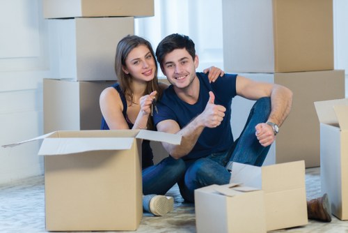 A young couple sitting on a light-colored carpeted floor inside a home surrounded by several cardboard moving boxes, some open and some closed, indicating a house relocation in progress. The woman has long brown hair and is wearing a sleeveless navy blue top, smiling and giving a thumbs-up gesture. The man has short dark hair, is dressed in a dark T-shirt and jeans, and is also smiling while giving a thumbs-up. Behind them, the background features a white wall with natural daylight coming through a window, typical of a residential interior in Croydon. The scene captures the moment of packing or unpacking, with relaxed and positive expressions, reflecting a professional removals service by Croydon Man with Van.