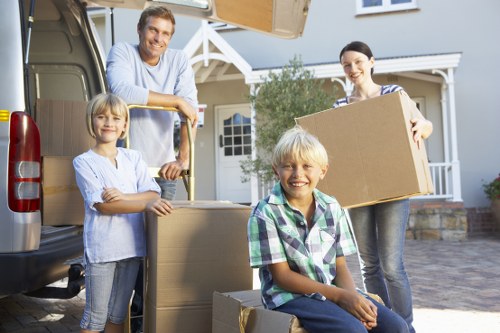 A family of four, including two children, are outdoors during daylight hours, loading or unloading a large cardboard box from a grey moving van parked in front of a residential property in Croydon. The father, wearing a light blue shirt, is standing at the back of the van, holding onto the edge of the open sliding door. The mother, dressed in a striped shirt, is standing on the driveway holding a large cardboard box in front of her. A young girl with blonde hair, wearing a white top and jeans, stands on the left side of the image, assisting with the move. A boy with blonde hair, wearing a checkered shirt, is seated on a cardboard box in the foreground, smiling towards the camera. The background features the front of a house with a porch, white railings, and a well-maintained garden, suggesting a typical residential setting in Croydon. The scene captures a typical house removal day with household furniture, cardboard boxes, and personal belongings being prepared for transit, illustrating the services of Croydon Man with Van in local house removals.