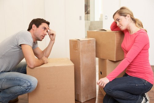 A young man and woman are indoors, surrounded by several cardboard moving boxes of various sizes, some stacked neatly on the floor in a residential setting with neutral walls and a staircase in the background. The man, dressed in a light grey t-shirt and blue jeans, is sitting on the floor, resting his head on his hand, appearing contemplative or slightly stressed. The woman, wearing a bright pink long-sleeve top and dark jeans, is sitting on a low step or platform, leaning on her arm, with a relaxed expression. The scene suggests they are in the process of packing or preparing for a house move, typical of a household in Croydon or nearby. The environment is well-lit with natural or indoor lighting, capturing the typical atmosphere of a household moving day, emphasizing the presence of moving supplies and the emotional state commonly associated with such activities, aligning with services offered by Croydon Man with Van in the Croydon area.