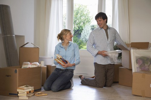 A spacious, well-lit living room with large windows revealing a green garden outside. Two individuals, a woman and a man, are surrounded by unpacked cardboard boxes filled with household items. The woman, with blonde hair tied back, is kneeling on the wooden floor and holding a stack of books with colorful covers. She is dressed in a light blue shirt and dark jeans. The man, with short dark hair, is standing and wearing a striped dress shirt and grey trousers. He is looking down at an open cardboard box containing more household items, such as plastic bags and packing paper. The scene captures a moment of unpacking during a house move facilitated by Croydon Man with Van, a local removals service near Croydon. The room has a neutral-toned wall and a window allowing natural daylight to illuminate the scene, emphasizing the casual, organized atmosphere of settling into a new home after a house relocation within the area.