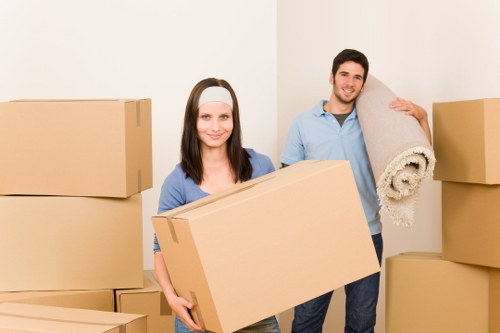 A young woman with long dark hair wearing a light blue shirt and a white headband stands amidst stacked cardboard moving boxes in a bright, plain white room, holding a large rectangular box. Beside her, a young man with short brown hair, dressed in a light blue shirt and dark trousers, carries a rolled-up beige rug over his shoulder, smiling at the camera. The boxes vary in size and are arranged around them, some stacked on the floor while others are supported against the wall. The environment appears to be a residential interior, likely during a house move, with the scene capturing a moment of packing or unpacking. The lighting is soft and natural, highlighting the neutral tones of the boxes and their casual clothing, suggesting a professional yet friendly house removals operation typical in the Croydon area.