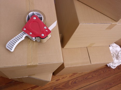 A close-up image showing a cardboard moving box on a wooden floor, with an orange and white tape dispenser placed on top of the box. The box appears to be partially opened or sealed, with additional cardboard boxes stacked nearby. The scene suggests a household environment, likely an interior space in a residential property, possibly during a house move or packing process. The tape dispenser is designed for sealing parcels, featuring a handle and wheel mechanism, and its bright orange color contrasts with the brown cardboard and polished wooden flooring. The overall setting indicates the logistics of packing or unpacking in a house, which is typical of house removals services in Croydon or surrounding areas in South London. This detail subtly supports local SEO for Croydon Man with Van and their specialising in removals services in the local region.
