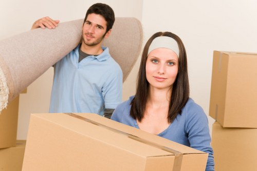 A young man with dark hair and a beard, wearing a light blue polo shirt, is standing behind a woman with long brown hair and fair skin, who is wearing a grey headband and a blue top. The man is lifting a large, light grey upholstered sofa or armchair, holding it on his shoulder with one hand. In the foreground, the woman is holding a medium-sized cardboard box, and there are additional cardboard boxes around them. The background shows a plain, neutral-colored wall, suggesting an indoor residential space, typical of a house or flat in Croydon. The lighting is bright and even, indicating a well-lit environment, possibly from natural daylight. The scene depicts a house removal or moving process, with items prepared for transportation. The overall setting appears organized and professional, consistent with services offered by Croydon Man with Van in the house removals sector.