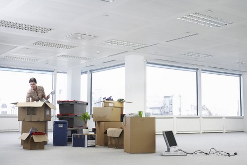 A professional woman in an office space with large windows, standing behind a collection of packed cardboard moving boxes of various sizes, some open revealing contents such as papers and small items. The boxes are stacked and scattered across the light-colored floor, with some placed on plastic crates for stability. A few boxes are marked or sealed with packing tape, and the woman appears to be preparing or organizing the belongings. The office is illuminated by natural daylight streaming through the windows, which offer a view of a cityscape with buildings in the background. The environment features a modern ceiling with recessed lighting panels and minimal furnishings, emphasizing a typical urban office setting in Croydon or surrounding areas, with no visible furniture aside from the boxes and a small black laptop on the floor, connected to a power cable. This scene illustrates steps involved in office removals or professional moving services provided by Croydon Man with Van, specializing in house and commercial relocations.