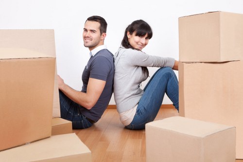 A young man and woman, sitting back-to-back on a light wooden floor in a bright, indoor space, surrounded by several cardboard boxes of varying sizes. The man, on the left, has short dark hair, a light beard, and is wearing a grey T-shirt and dark jeans, smiling as he points to a box in front of him. The woman, on the right, has dark hair tied in a ponytail and is dressed in a grey hoodie and blue jeans, smiling as she leans against a stack of two large cardboard boxes. The scene suggests a house move or packing activity, with the boxes ready for transportation or storage. The background is plain white, emphasizing the household setting, and the overall atmosphere appears organized and calm, typical of a professional house removals or relocation process in Croydon or the surrounding area, in line with Croydon Man with Van’s services focused on sustainable and efficient moves within the local community.