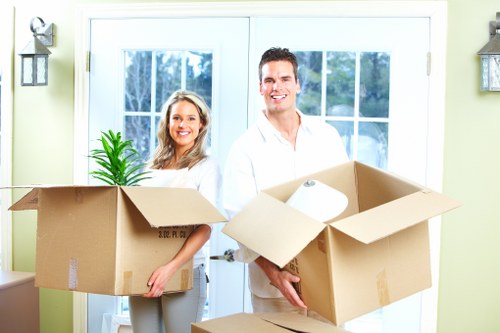 A man and woman, both smiling, stand indoors in front of a large glass door with white framing, holding open two cardboard boxes filled with household items during a house move. The woman, positioned on the left, has shoulder-length blonde hair and wears a white top, holding a box containing a small potted plant with vibrant green leaves. The man, on the right, has short dark hair, and is dressed in a white shirt, carrying a box that contains a few rolls of paper or packing material. Behind them, the light-colored interior features a pair of matching wall-mounted lantern-style light fixtures on either side of the door, enhancing a welcoming residential atmosphere. The boxes are made of brown corrugated cardboard, showing signs of being used for packing, with flaps open to reveal the contents. Natural daylight filters through the glass panels, illuminating the scene, which reflects a typical residential setting suitable for a local house removals service in Croydon. The scene clearly illustrates preparation for moving house, with a focus on household furniture and packing supplies, fitting within the context of house removals and local relocation services.