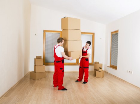Two removalists from Croydon Man with Van, dressed in red uniform with black accents, are inside a bright, empty room with light-colored wooden flooring and white walls. They are stacking a large cardboard box on top of several other boxes already arranged vertically, forming a tall column. Additional cardboard boxes are visible on the floor near the window and against the wall, some of which are closed and others partially open or stacked. The room has three large windows with wooden frames; one directly behind the removalists and another on the right side, allowing natural light to illuminate the space. The environment appears to be a residential property, preparing for a house removal or relocation in Croydon, with no furniture present. The neutral setting features sparse furnishings, focusing on the packing process essential for professional removals services in the local area.