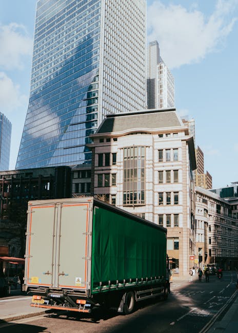 The image depicts a scene involving home relocation or furniture transport. Inside a property's doorway, a mover is seen carrying a cardboard box wrapped in protective plastic wrap, positioned on the