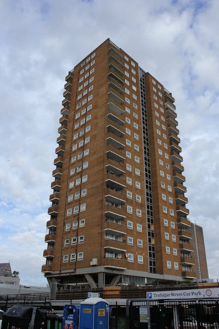 Photograph of the upper floors of a high-rise residential building during daytime, showing multiple rows of windows and balconies with some laundry hanging outside. A blue and white modern apartment b